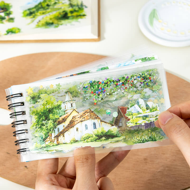 HandsholdingaclearPETstickersheetfeaturingacountrysidecottageandfloweringgreenery