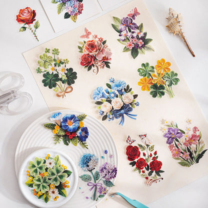 A flat-lay display of colorful floral and botanical illustrations printed on sheets and stickers, arranged on a white surface with decorative props including a seashell, clear clips, and a small dish.