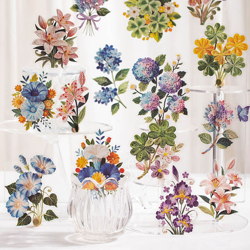 A collection of colorful floral illustrations arranged on clear glass display stands and vases against a soft white background. The artwork features watercolor-style flowers including lilies, hydrangeas, clovers, bluebells, and purple blossoms.
