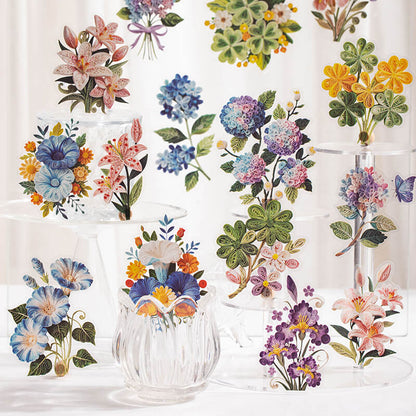 A collection of colorful floral illustrations arranged on clear glass display stands and vases against a soft white background. The artwork features watercolor-style flowers including lilies, hydrangeas, clovers, bluebells, and purple blossoms.