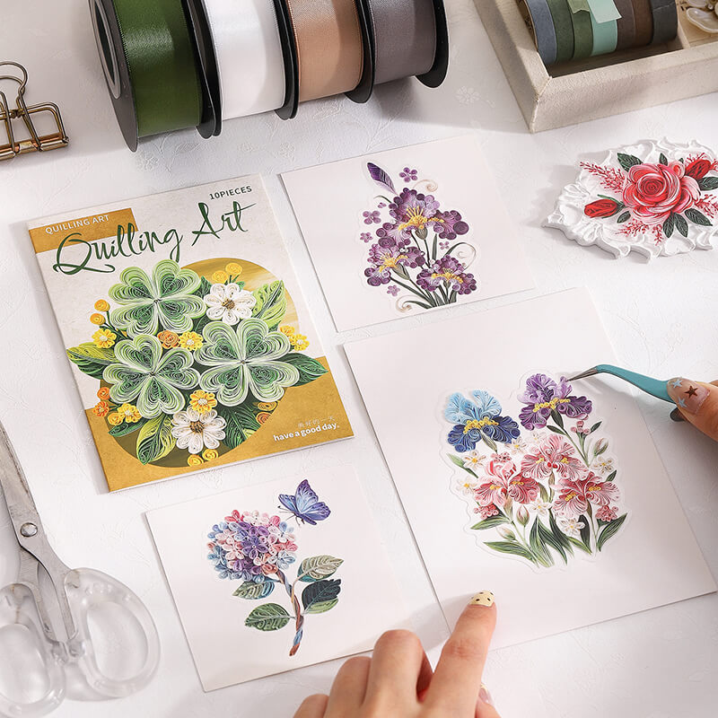 A styled flat-lay of floral sticker art supplies on a white surface, including four printed floral sticker sheets, a Quilling Art booklet, ribbon spools, scissors, and tweezers. A hand is placing a pink and purple flower sticker, with another hand holding a teal tool.