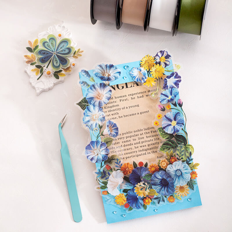 A decorated rectangular card or sticker sheet with a blue floral border and a central text panel, arranged on a white surface with a small turquoise tool, a floral embellishment, and ribbon spools nearby.