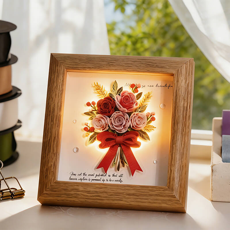 A square wooden shadow box candle holder featuring a warm glowing floral design with pink and red roses, green leaves, and a red ribbon, displayed on a windowsill with soft natural light.