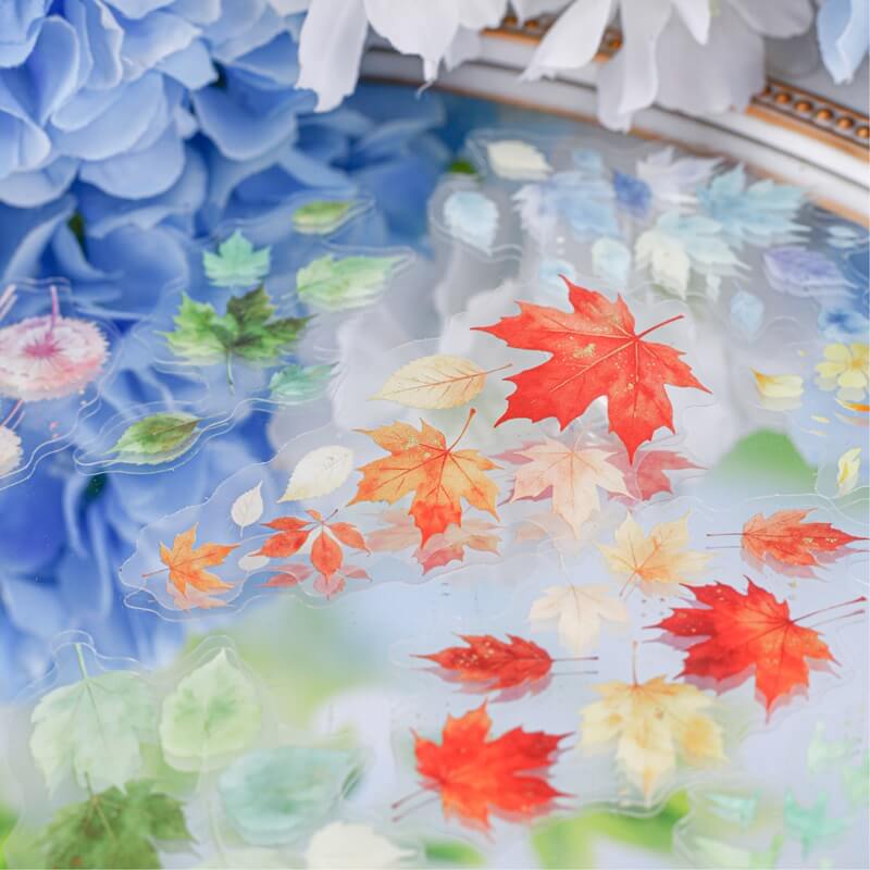 A close-up of a decorative tray or dish filled with colorful autumn leaf decals or stickers, including red, orange, yellow, and green maple leaves, layered over a soft blue floral background.