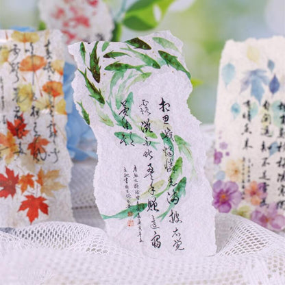 Three handmade paper cards with torn edges and Chinese calligraphy, each decorated with different watercolor patterns (red autumn leaves, green bamboo, and blue/purple flowers), arranged on a white mesh surface.